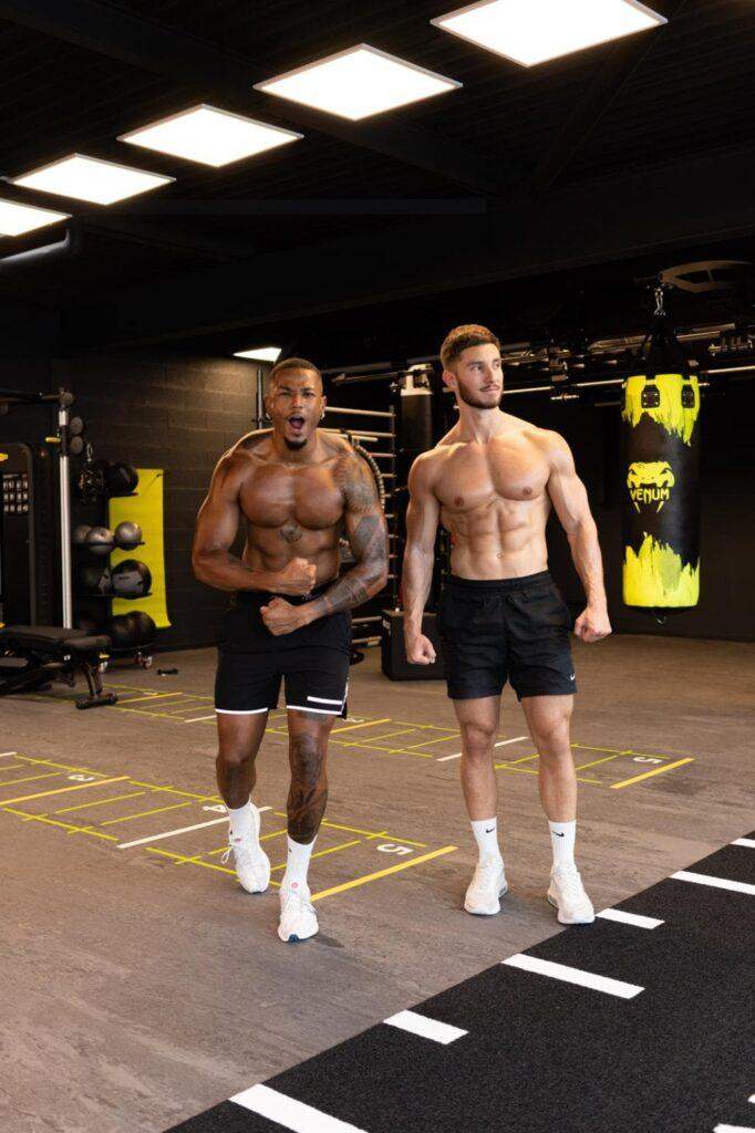 Deux hommes dans une salle de sport posant pour une photo, tous deux torse nu et fléchissant leurs muscles. Ils portent un short noir et des baskets blanches. Des équipements de gymnastique et un sac de boxe sont visibles en arrière-plan.