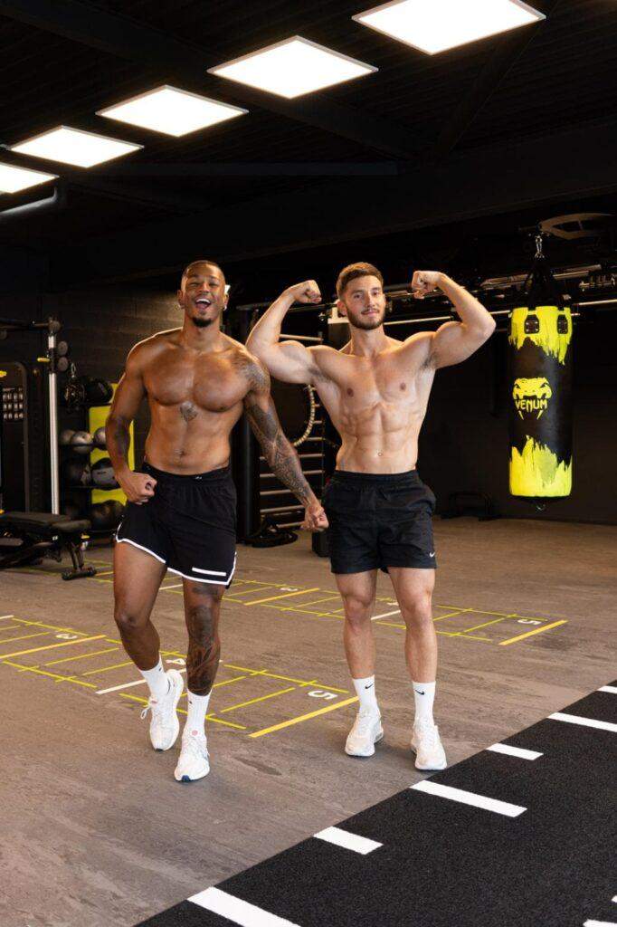 Deux hommes torse nu en short de sport posent dans une salle de sport, fléchissant leurs muscles et souriant. Des équipements d’exercice, dont un sac de boxe, sont visibles à l’arrière-plan.