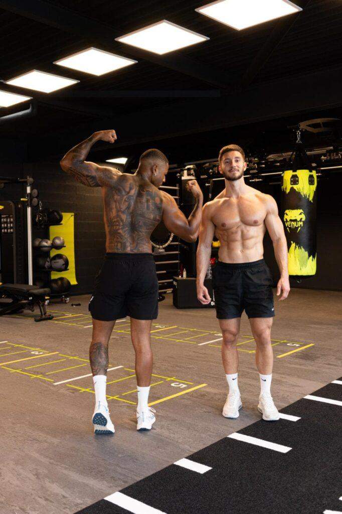 Deux hommes dans une salle de sport, l'un avec des tatouages dans le dos fléchissant le bras et l'autre debout, les mains sur les côtés. Tous deux sont torse nu et portent un short noir et des chaussures blanches. Les équipements de fitness sont en arrière-plan.